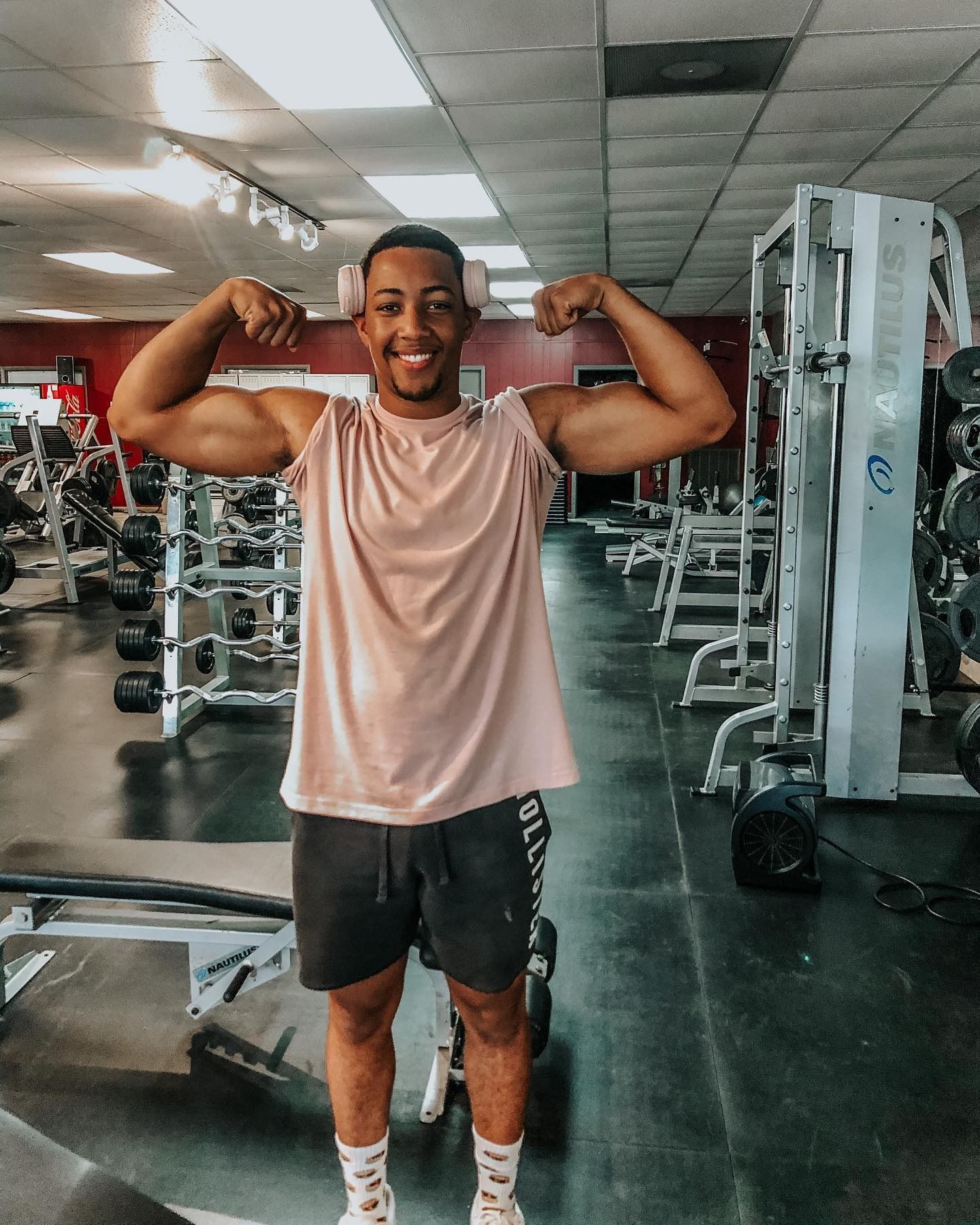 A smiling man flexes his biceps in a gym. He wears a pink shirt, black shorts, and headphones.