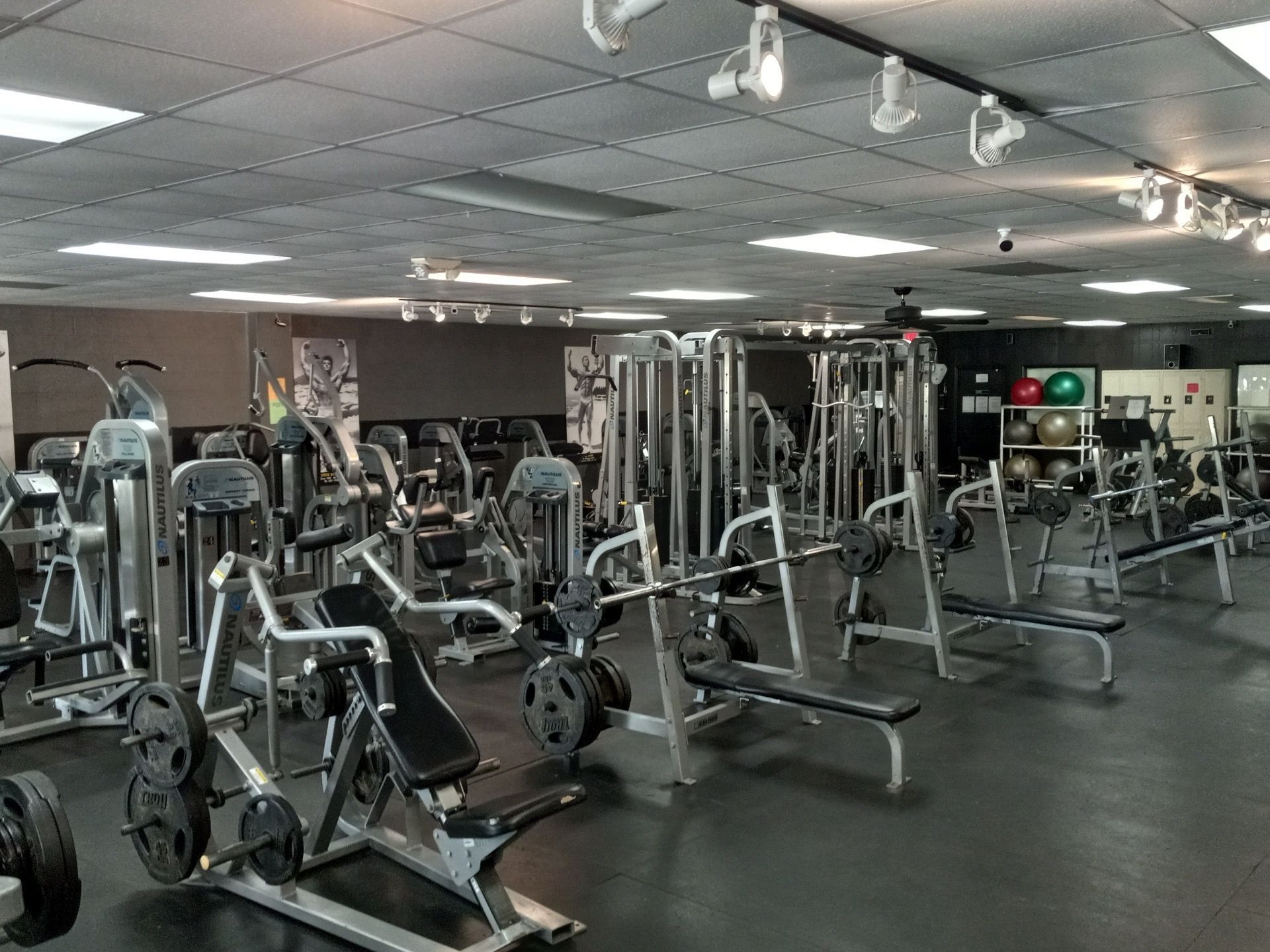 A fitness gym with various exercise machines and weights. Black flooring, gray ceiling and walls, and bright lights.