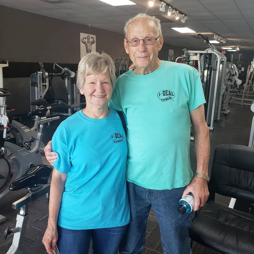 A senior couple smiles in a gym. They wear matching teal shirts. The man has his arm around the woman. Gym equipment is in the background.