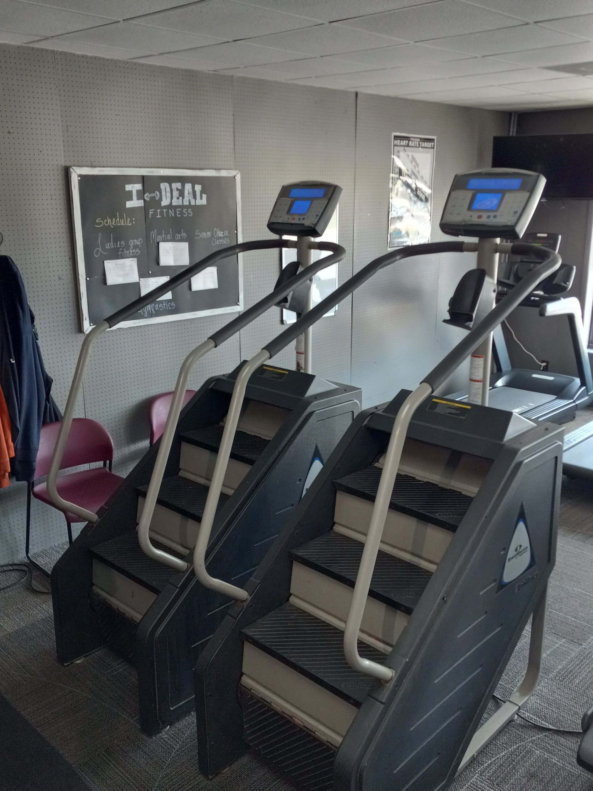Two stair-step machines in a gym. Dark gray machines with screens and handrails, set against a gray wall with a notice board.