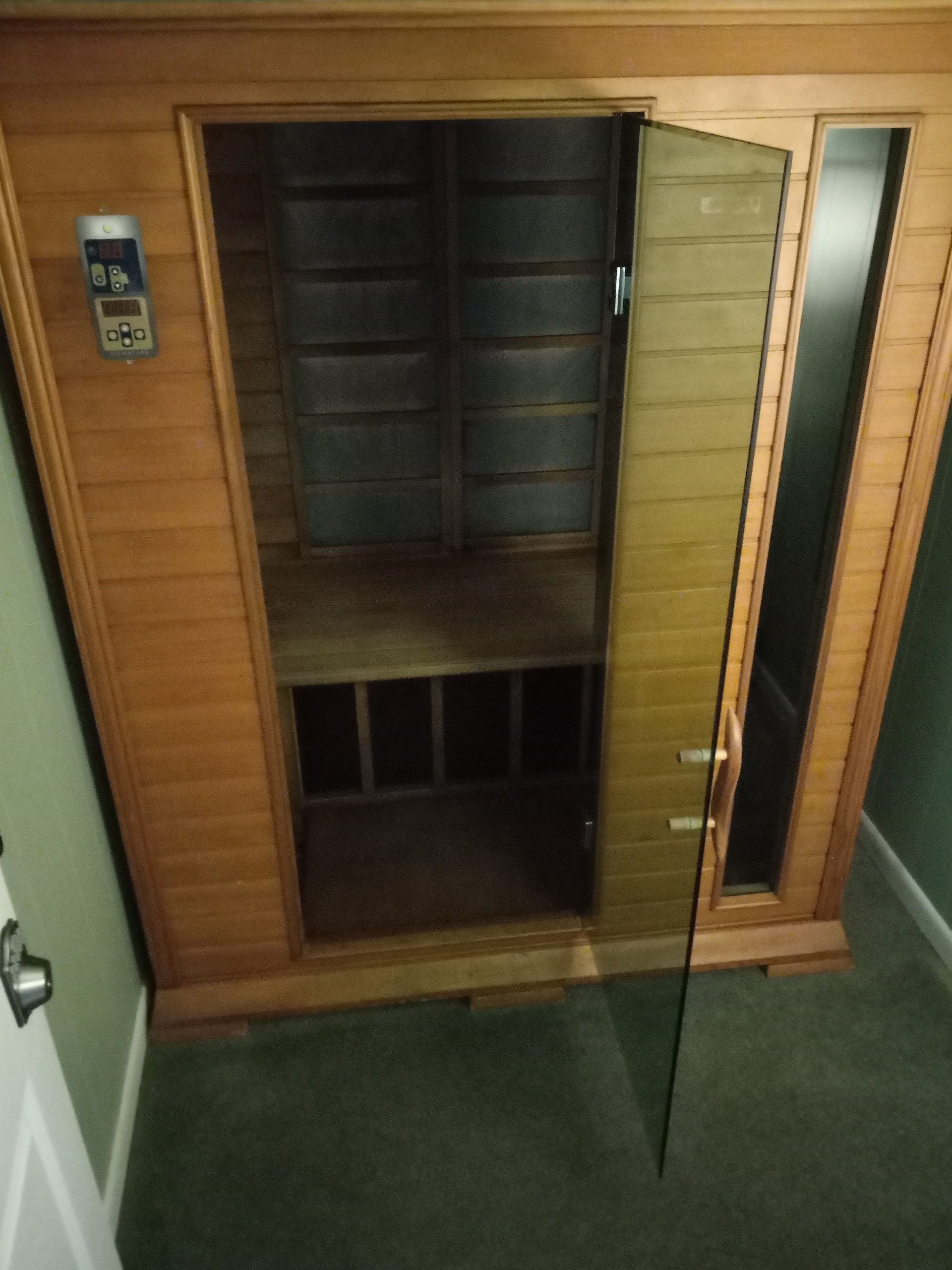 A wooden sauna with an open glass door, revealing the interior. The sauna is in a room with green carpet.