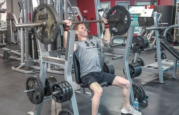 Man lifting weights on a seated shoulder press machine at a gym. He is wearing a grey shirt and black shorts.