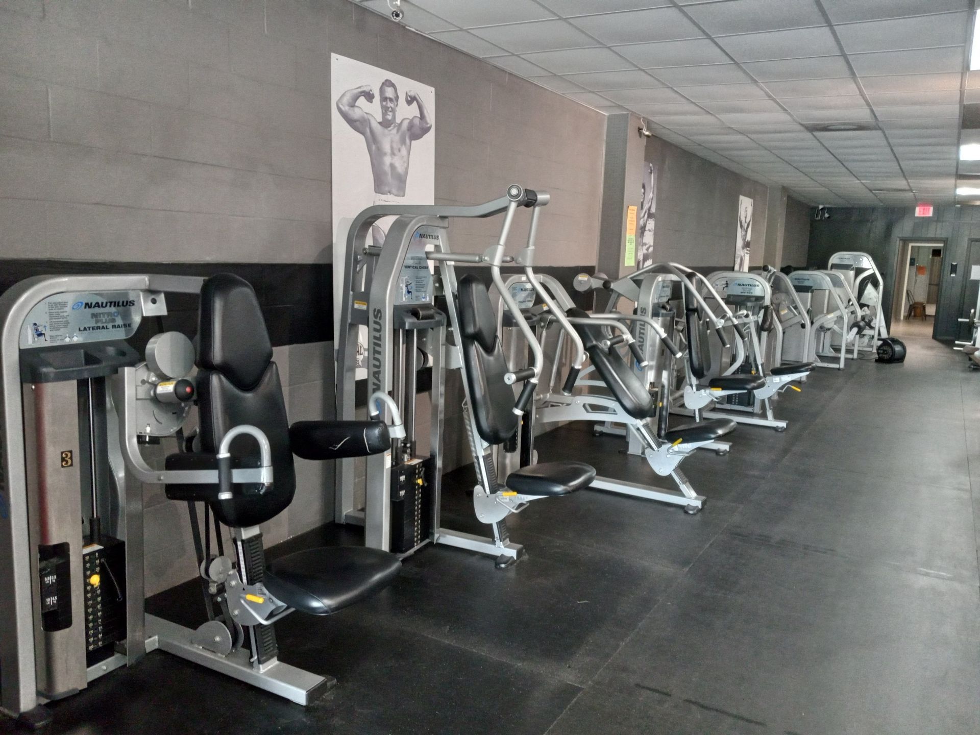 Row of exercise machines in a gym, including leg press, chest press, and shoulder press. Gray walls, black floor.