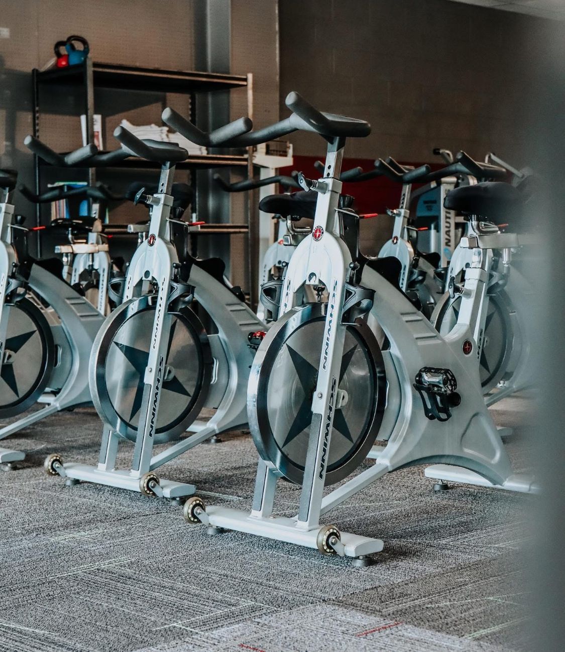 Row of white stationary bikes in a dimly lit gym with gray carpet.