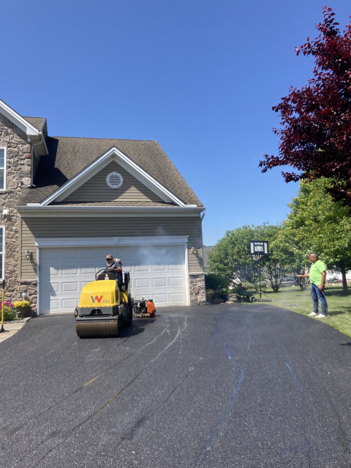 A man in a green shirt is standing in front of a house while a roller is working on the driveway.