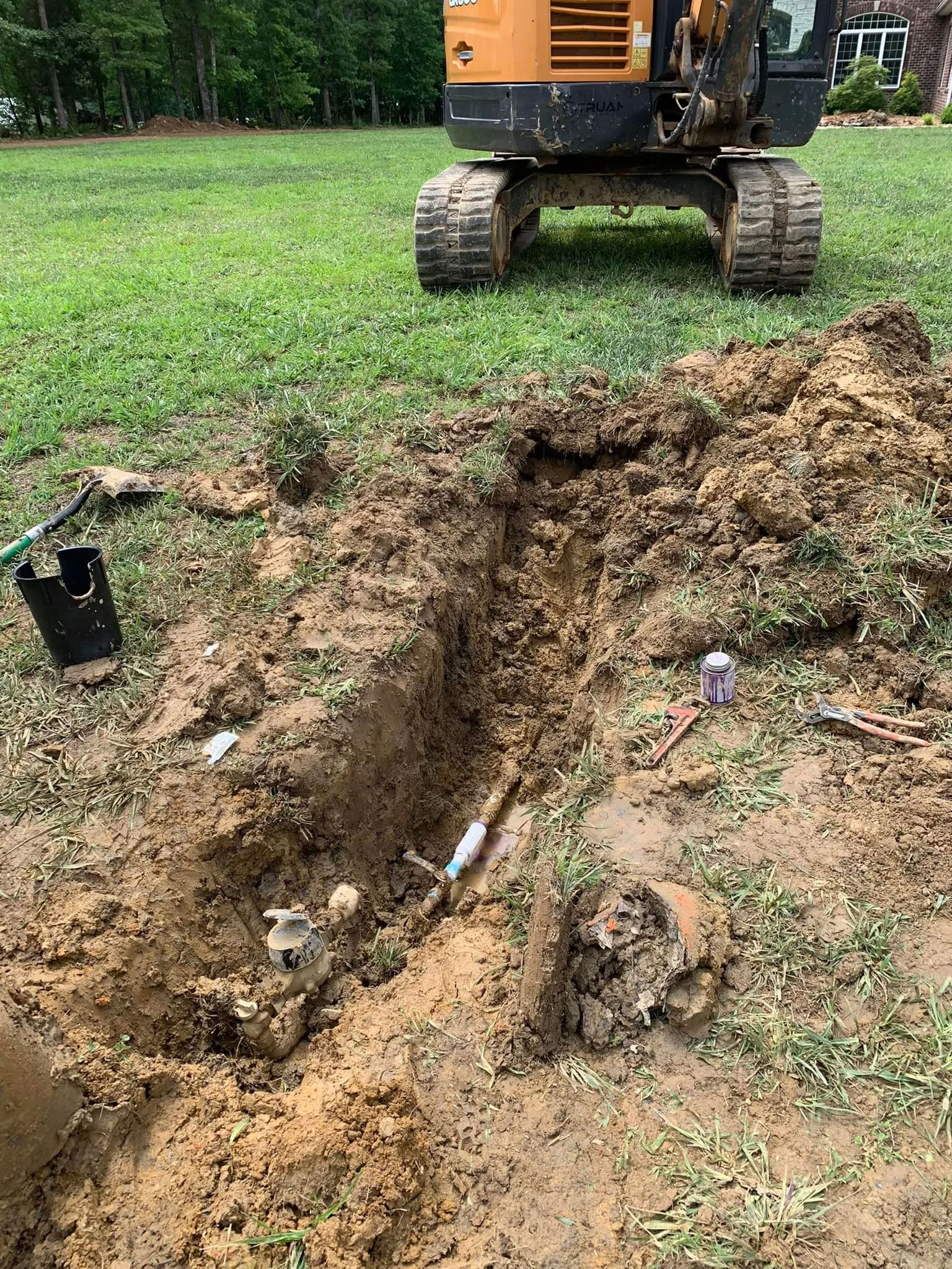 Excavator beside a muddy trench in a grassy yard, with exposed roots and dirt piled nearby