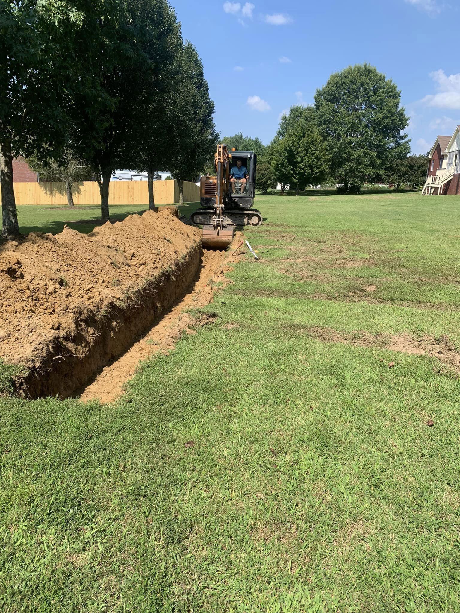 Excavator digging a long trench beside a line of trees in a grassy field on a sunny day