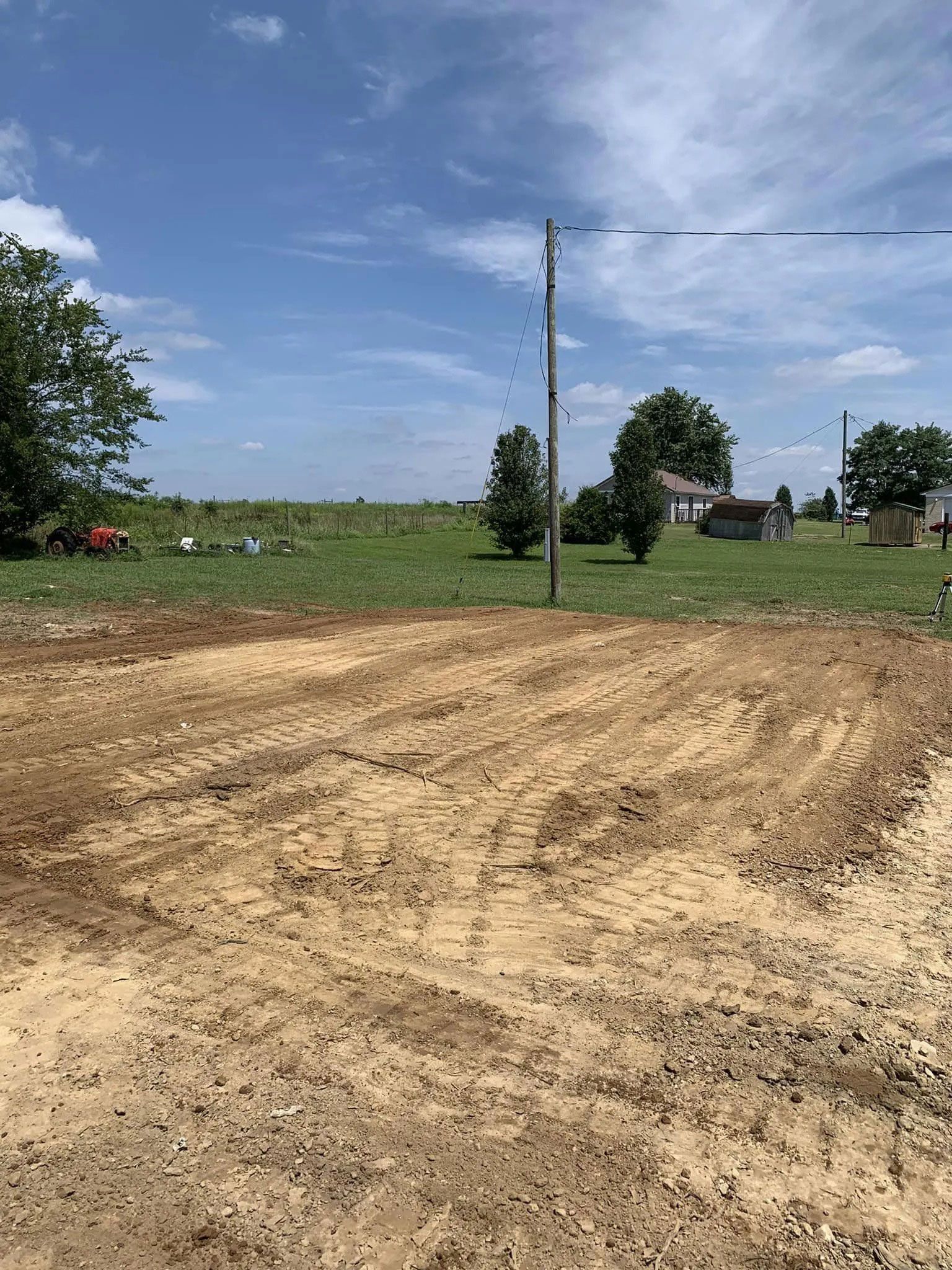 Freshly tilled dirt field with a utility pole, trees, and a farmhouse under a blue sky