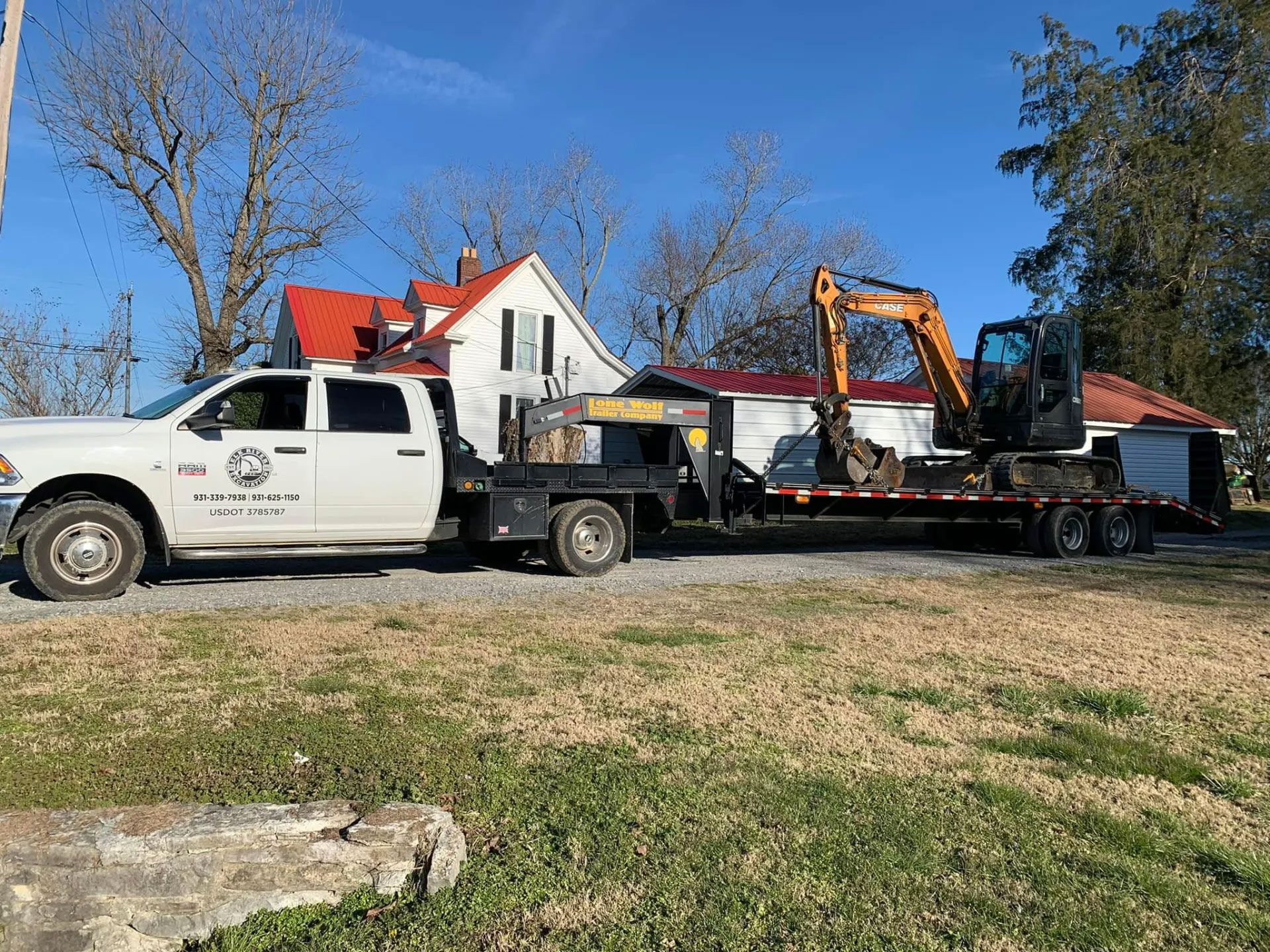 White work truck towing an excavator on a trailer in a yard, with a house and trees in the background