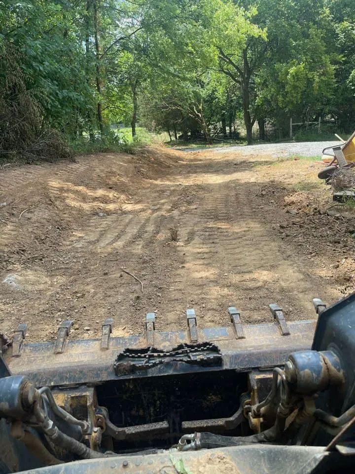 View from a loader bucket toward a dirt path through trees, with a pile of rocks on the right.