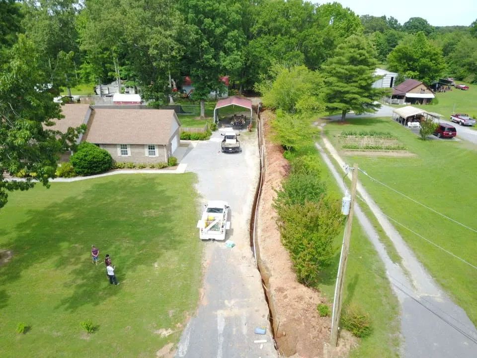 Aerial view of a rural road repair beside homes and a grassy field, with workers and vehicles below.