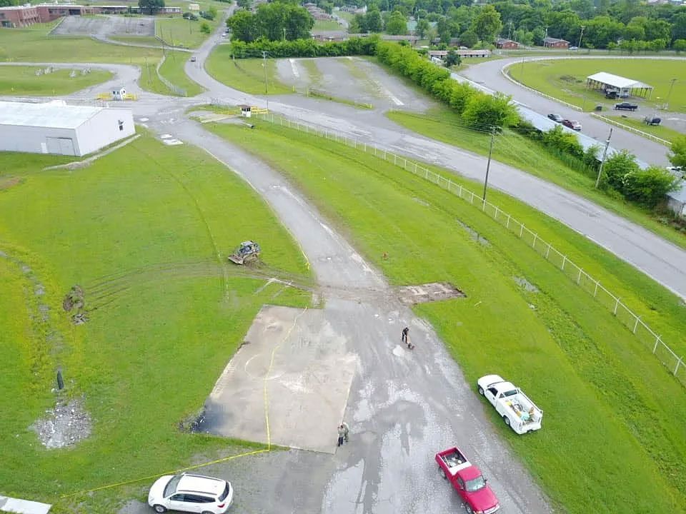 Aerial view of a grassy lot, paved road, and parked cars near a white building.