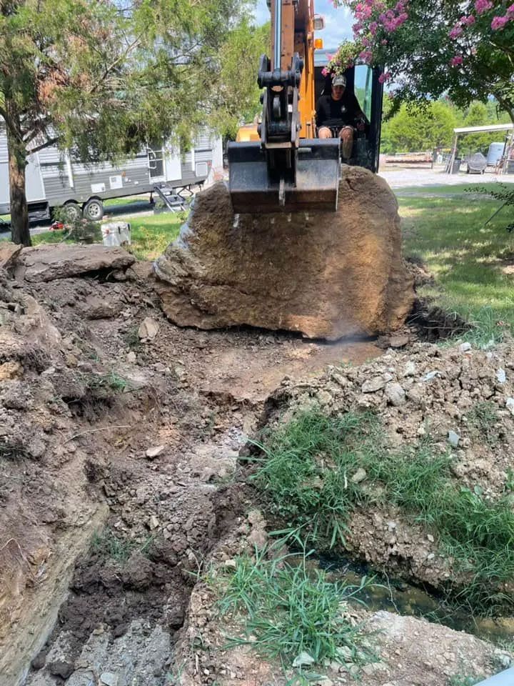 Excavator digging up a muddy yard, lifting a large clump of soil and roots.