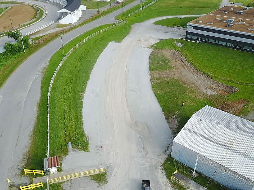 Aerial view of a rural road, grassy embankments, and farm buildings beside a gravel lot.