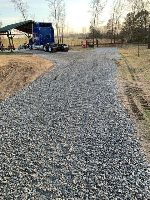 Gravel driveway being built beside a blue truck and covered structure in a rural yard.