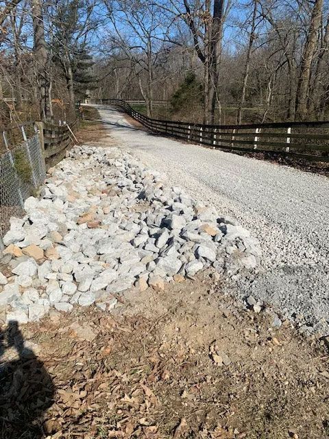 Gravel road with rock pile, lined by a fence and wooden guardrails in a wooded area