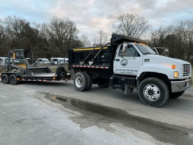 White dump truck towing a flatbed trailer with a skid steer in a parking lot