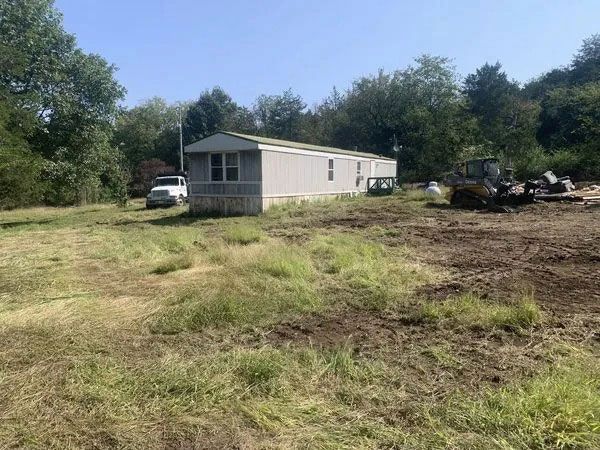 Mobile home on a grassy lot with a white truck and construction equipment nearby under a clear sky