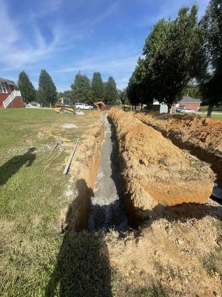 Trenched dirt ditch beside a grassy field under a blue sky, with trees and houses in the background