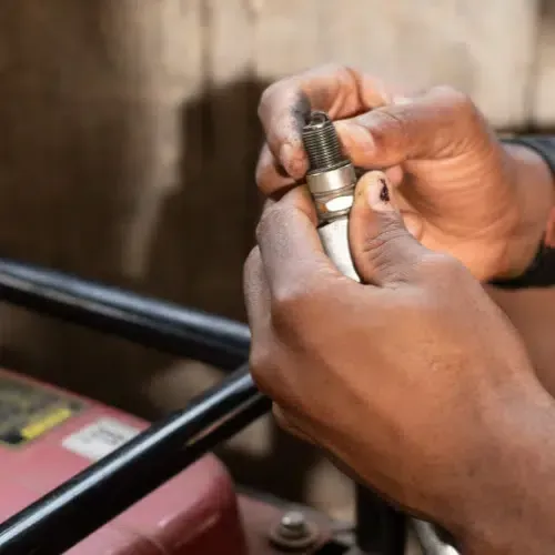 Close-up of hands holding and inspecting a used spark plug, with a red generator visible in the background.