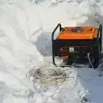 An orange portable generator and a coiled white extension cord sit on a thick layer of snow.