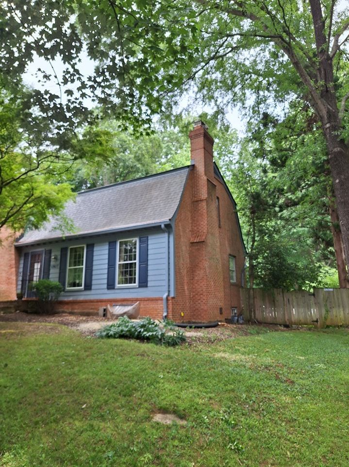 A small brick house with a blue siding and a chimney in the backyard surrounded by trees.