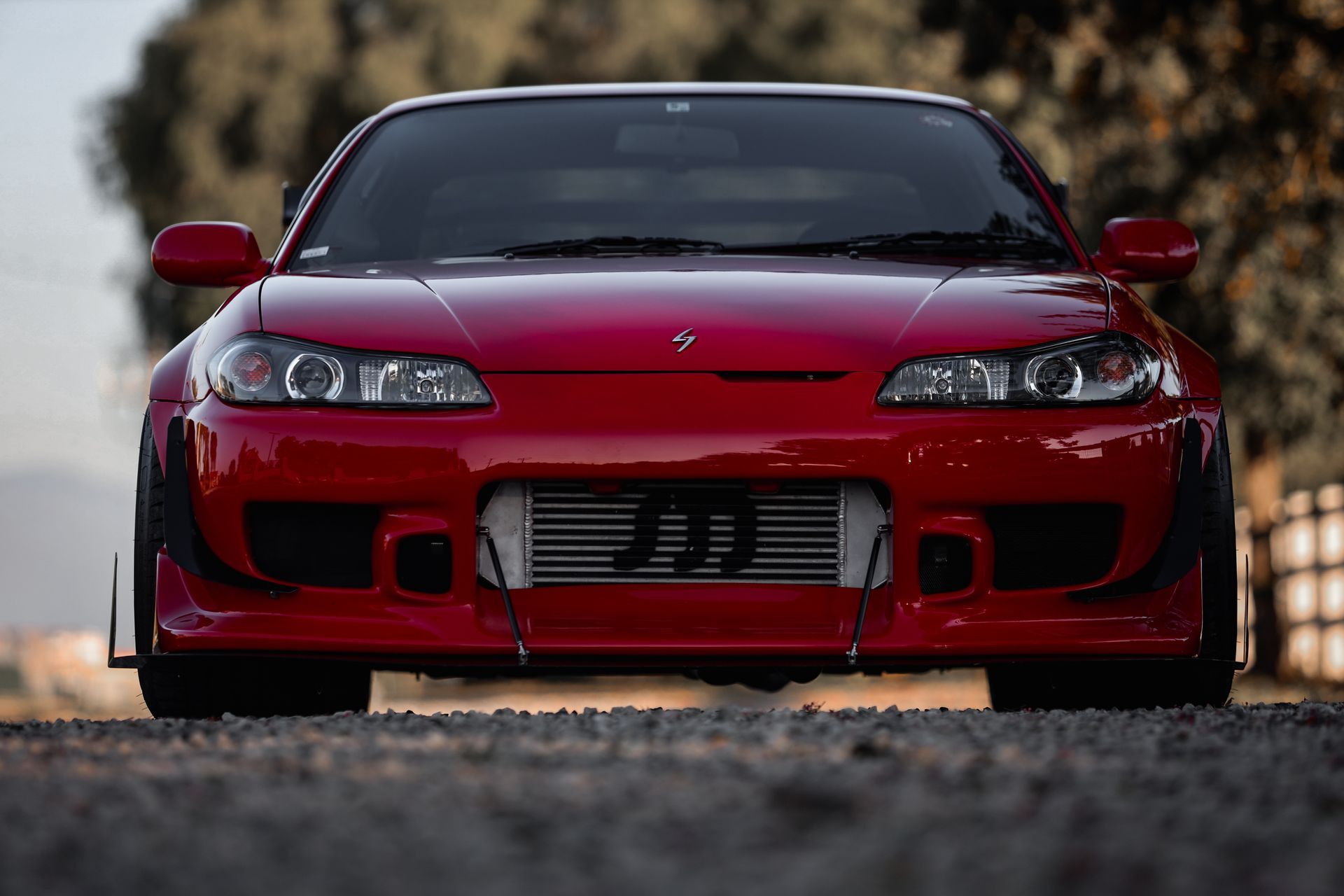 Red Nissan S15 sports car with widebody kit, parked on a gravel road.