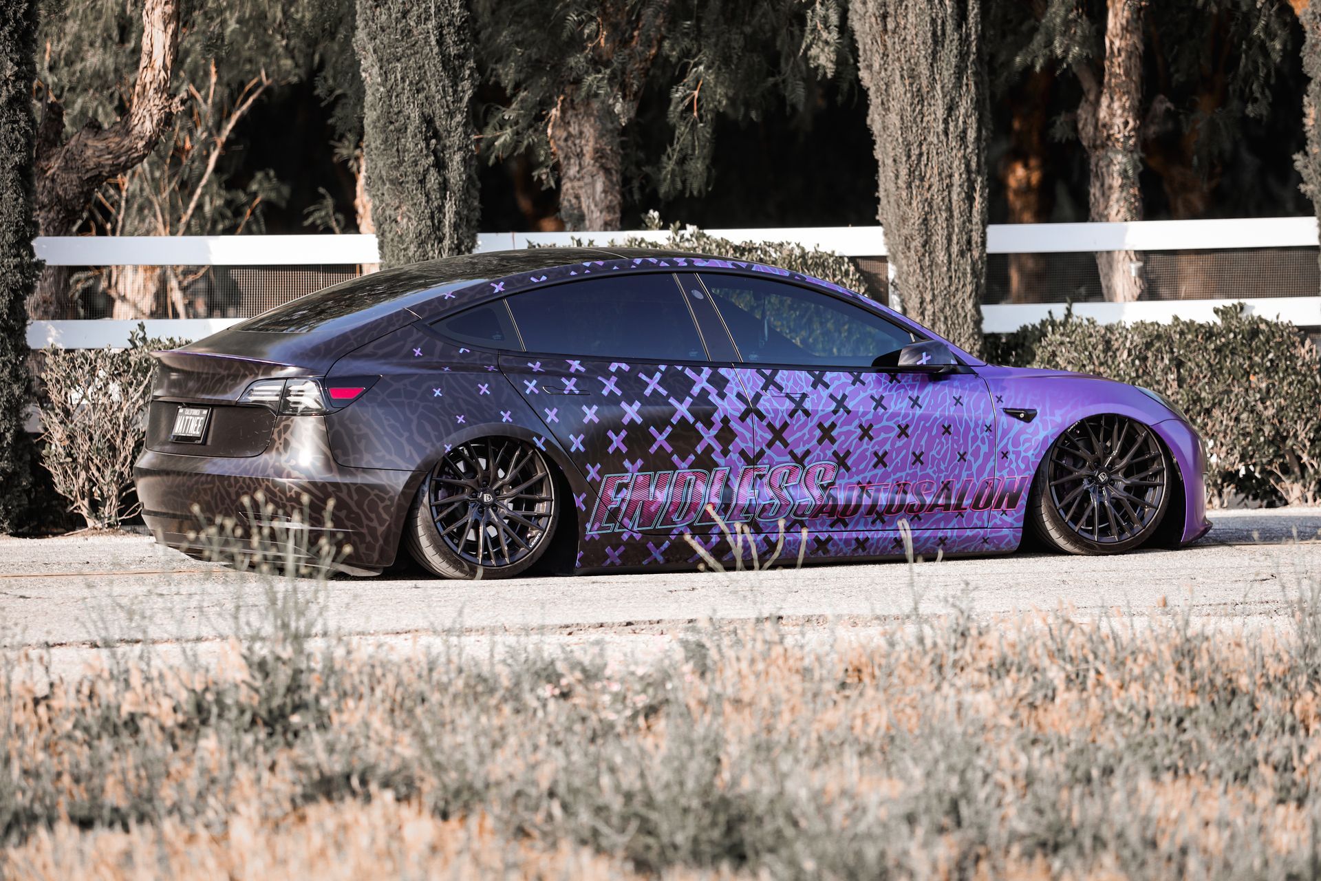 Purple-wrapped Tesla Model 3 car on a gravel road, lowered suspension, black wheels, against a background of trees and a white fence.