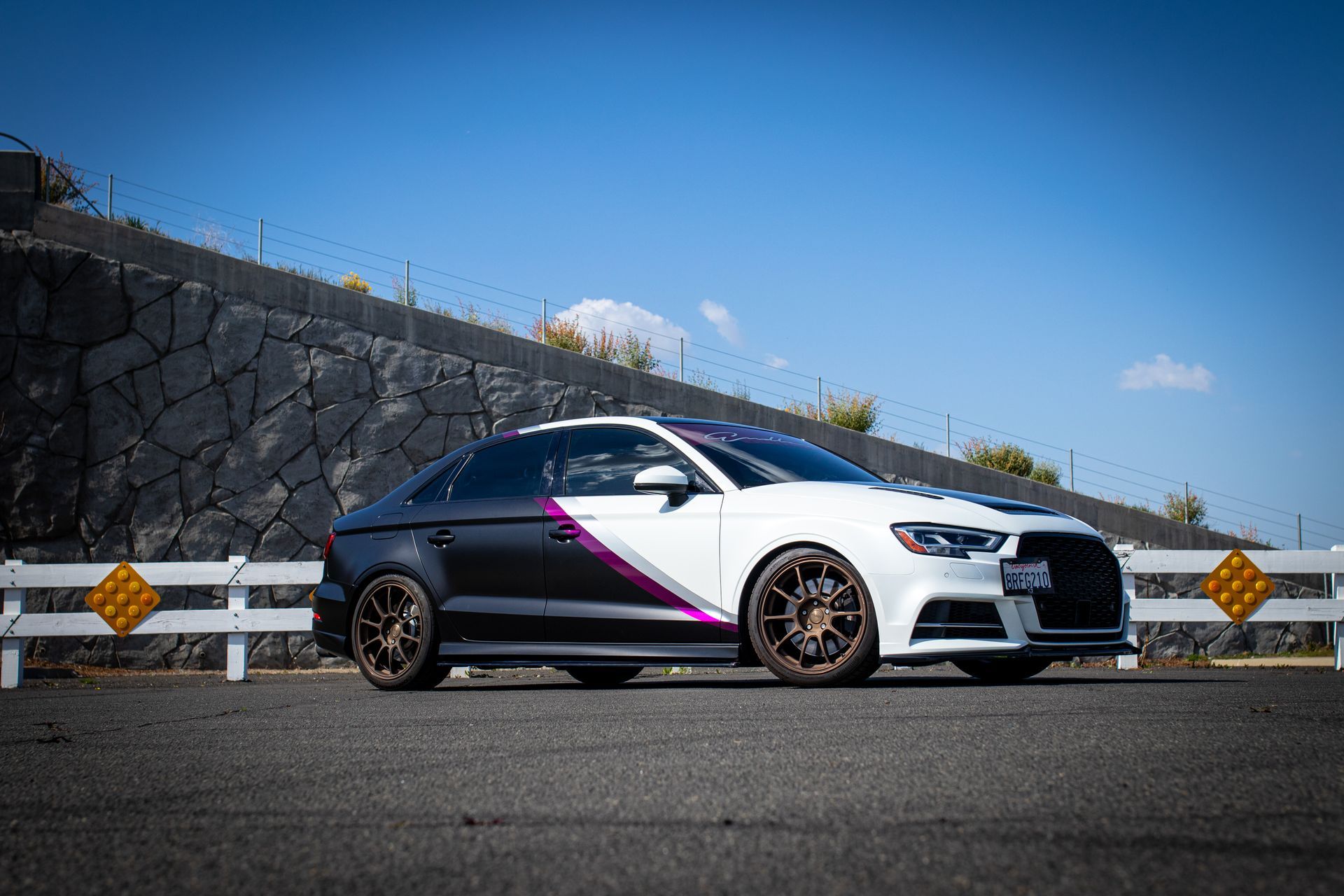 Customized black and white Audi sedan with bronze wheels parked on asphalt road near a stone wall.