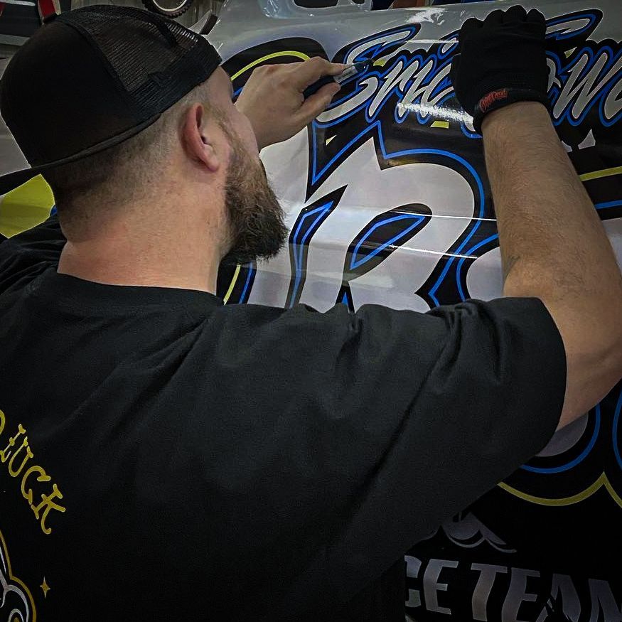 Man applying a decal to a car. He wears a black cap and gloves. The decal is blue, yellow, and white.