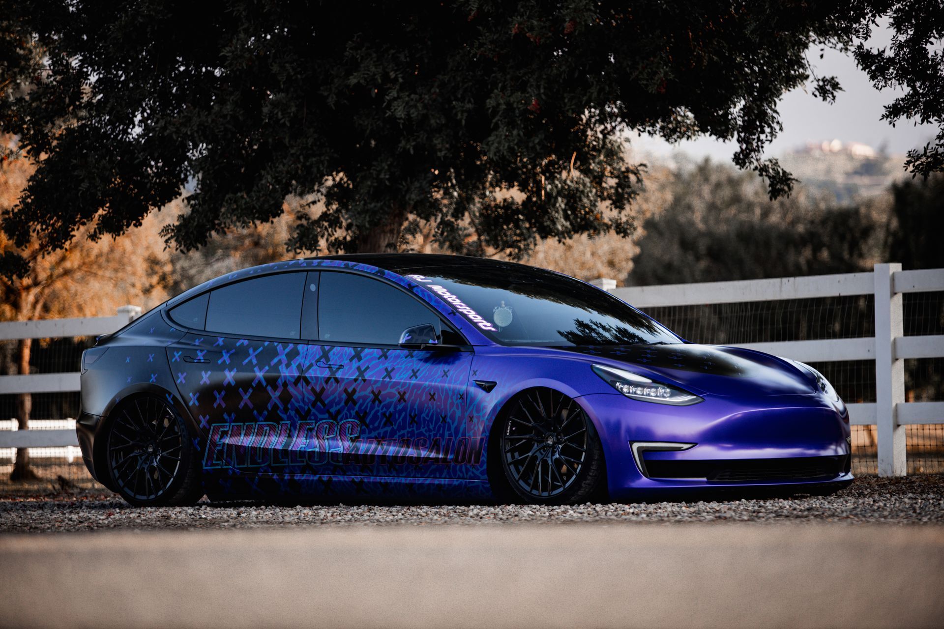 Modified Tesla Model 3 in purple with black wheels, parked in front of a white fence.