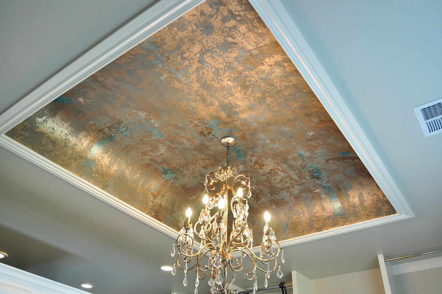 Ornate chandelier hanging below a textured copper-colored ceiling with turquoise accents, framed by white molding.
