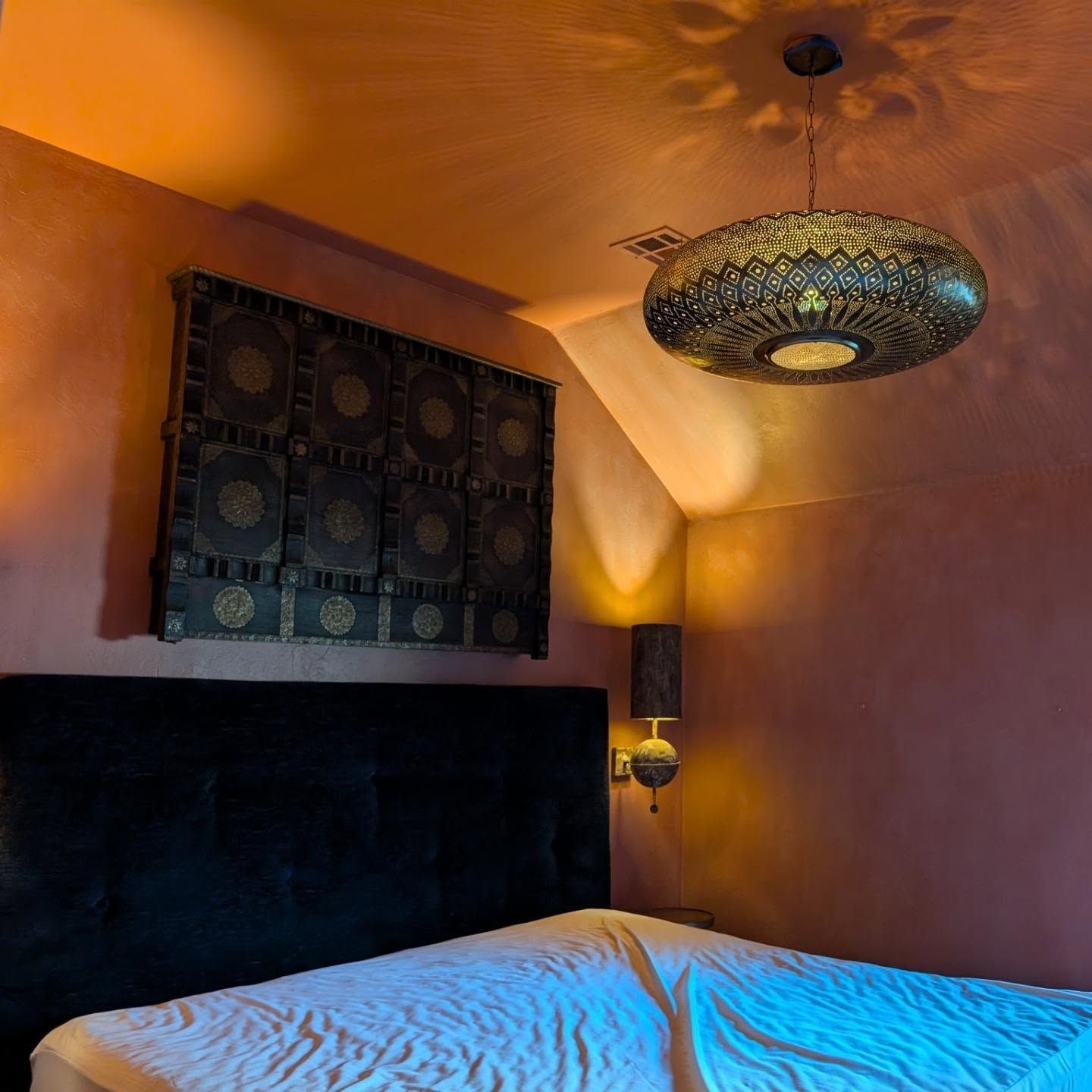 Bedroom with ornate ceiling light, wall art, and black headboard. Pink walls, warm lighting.