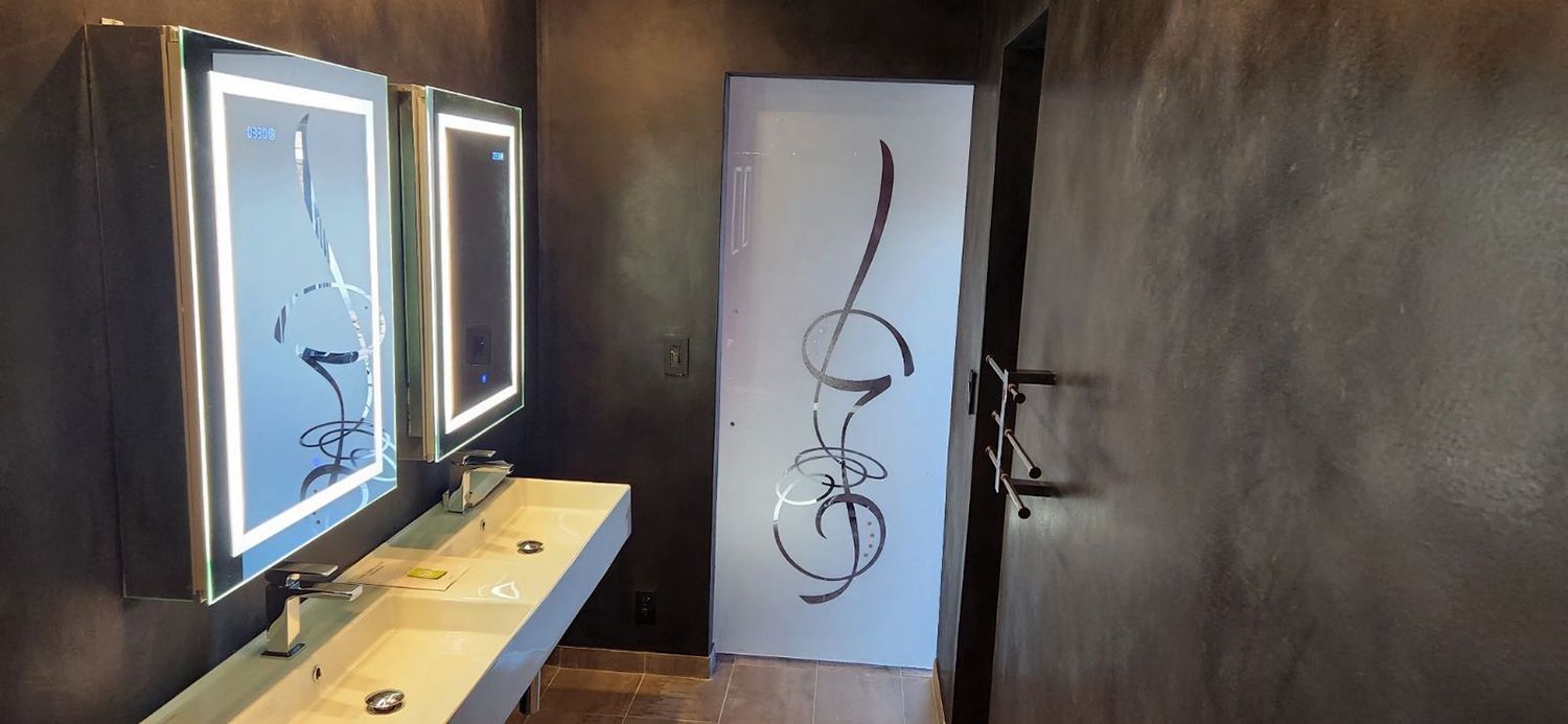 Bathroom with two illuminated mirrors, sink, and a decorative frosted glass door. Dark walls.