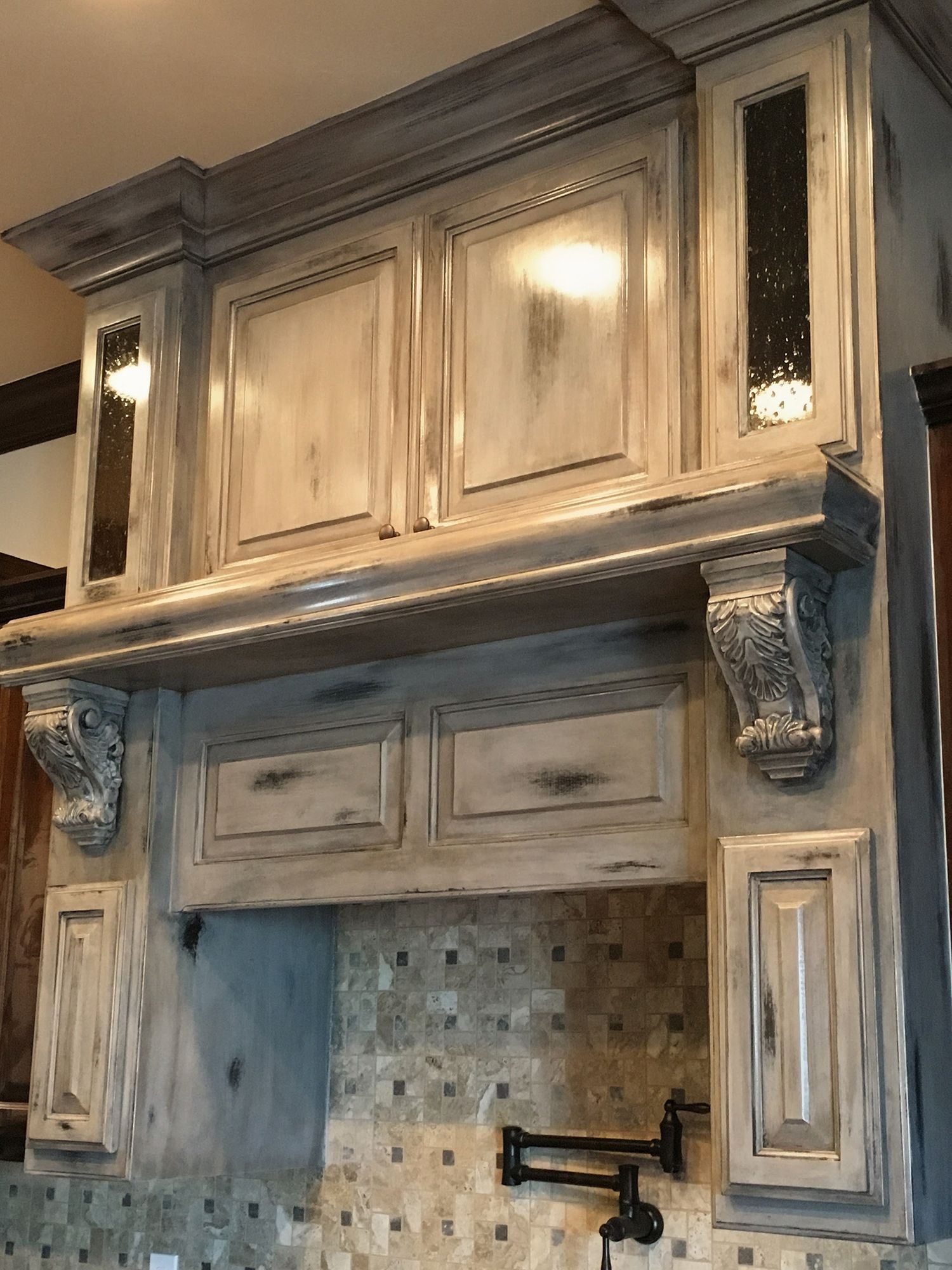 Gray, distressed kitchen range hood with ornate details, glass-front cabinets, and mosaic tile backsplash.