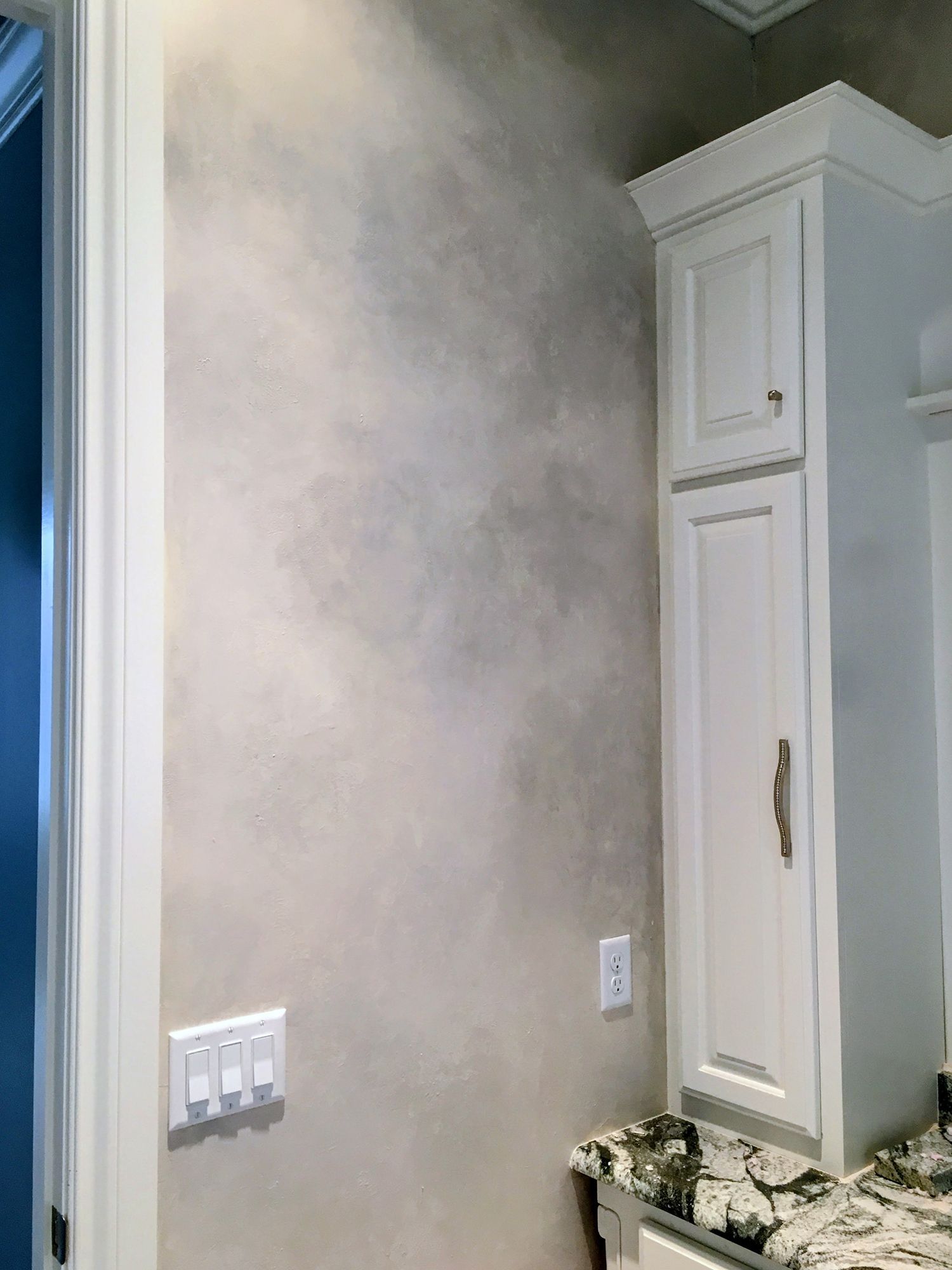 Wall with textured gray finish, white cabinets with granite countertop, light switches, and a doorway.