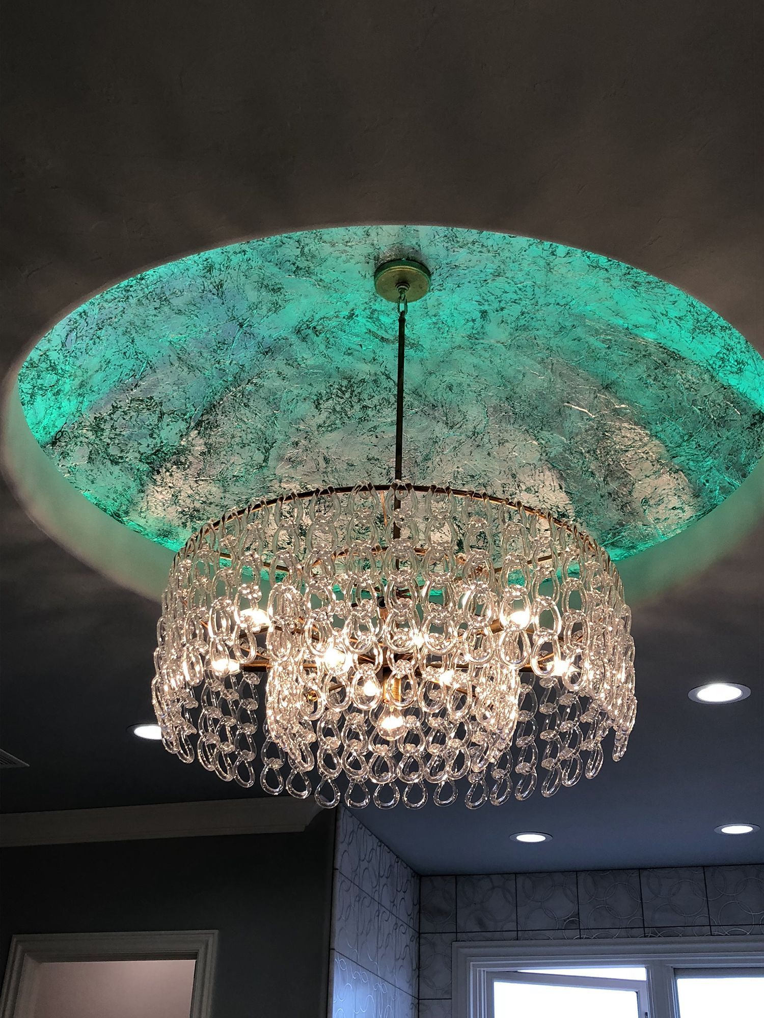 Crystal chandelier hanging from a green-lit recessed ceiling in a room.