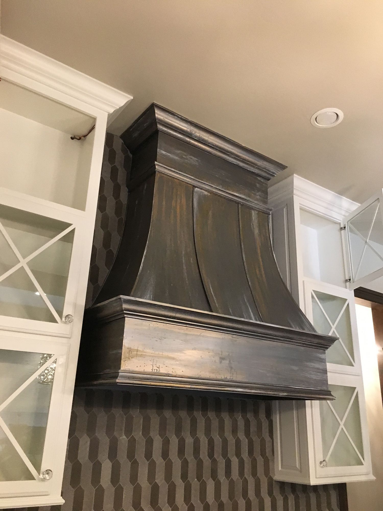 Metallic range hood above a stovetop, flanked by white cabinets, mounted on a mosaic tile wall.