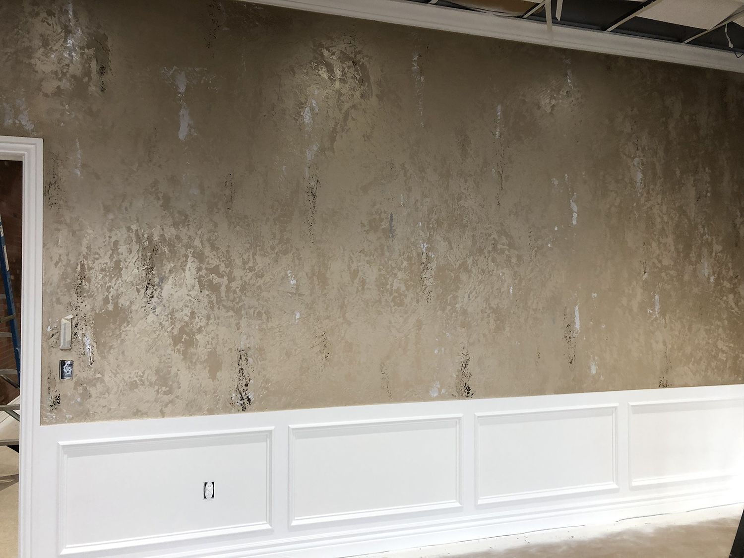Wall with textured tan and silver finish above white wainscoting. Electrical outlets and a doorway are visible.