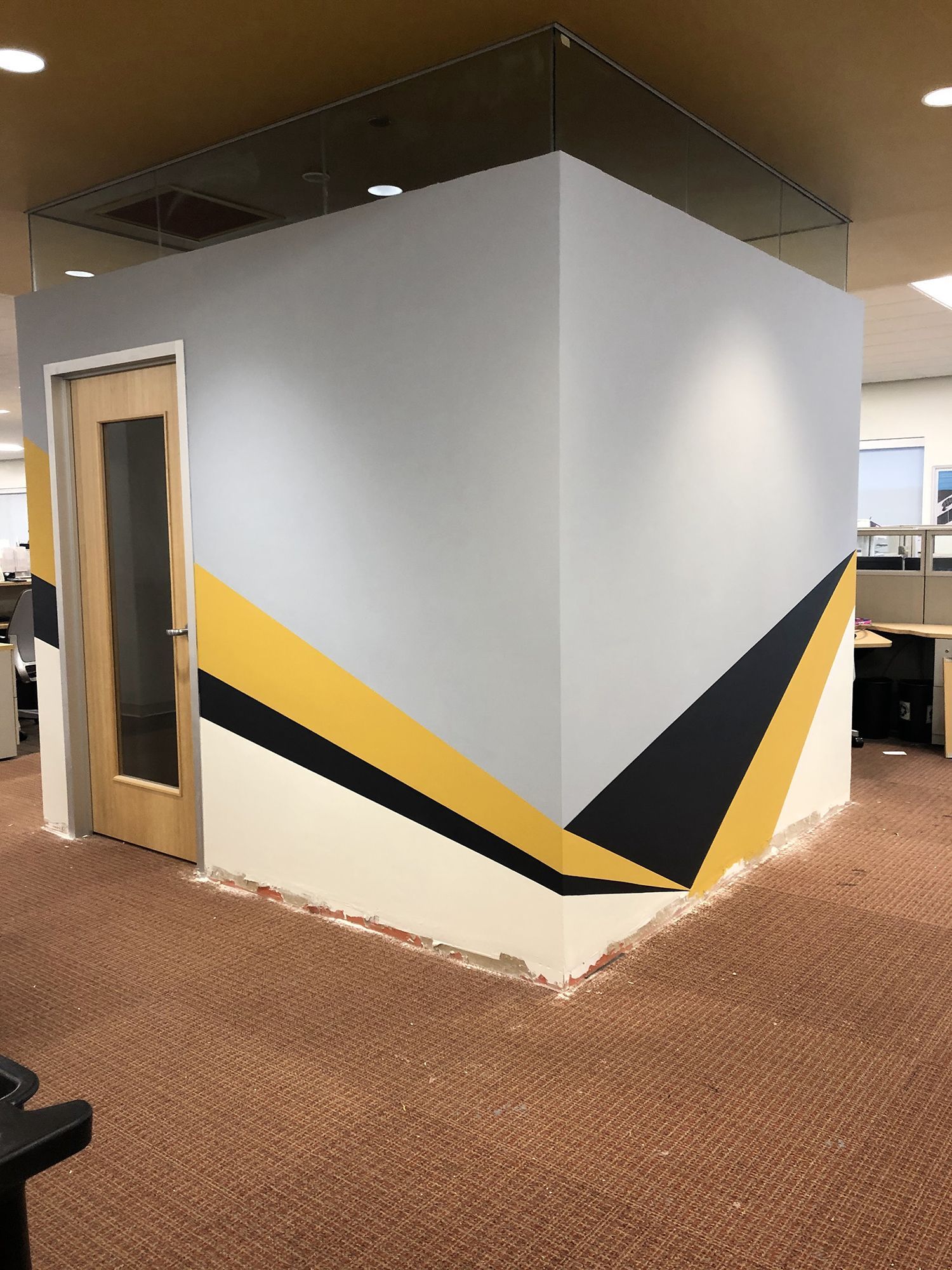 Gray office cubicle with yellow and black geometric design; wooden door; brown carpet.
