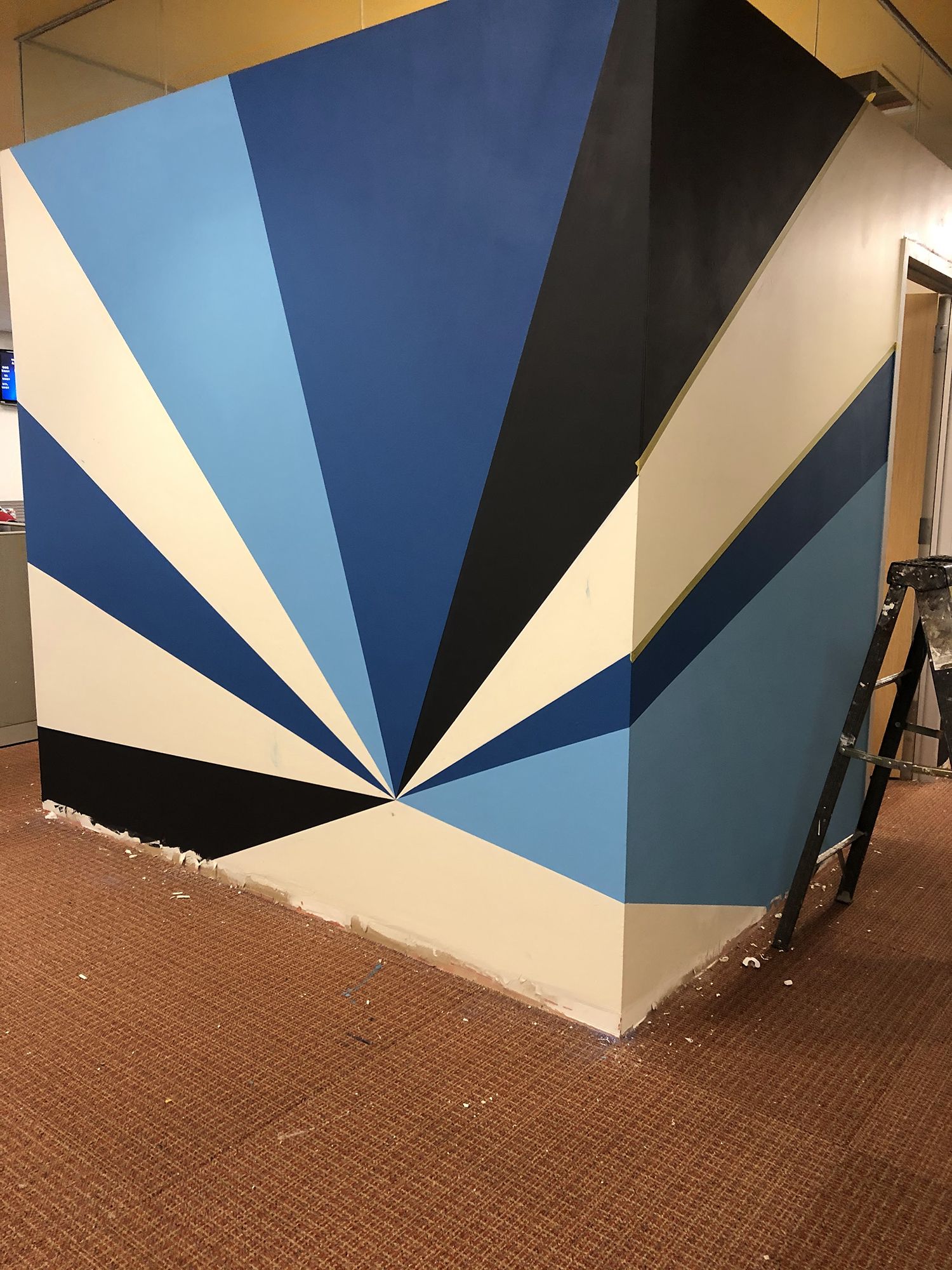 Geometric mural in shades of blue, black, and beige on a corner wall. A small step ladder stands nearby.
