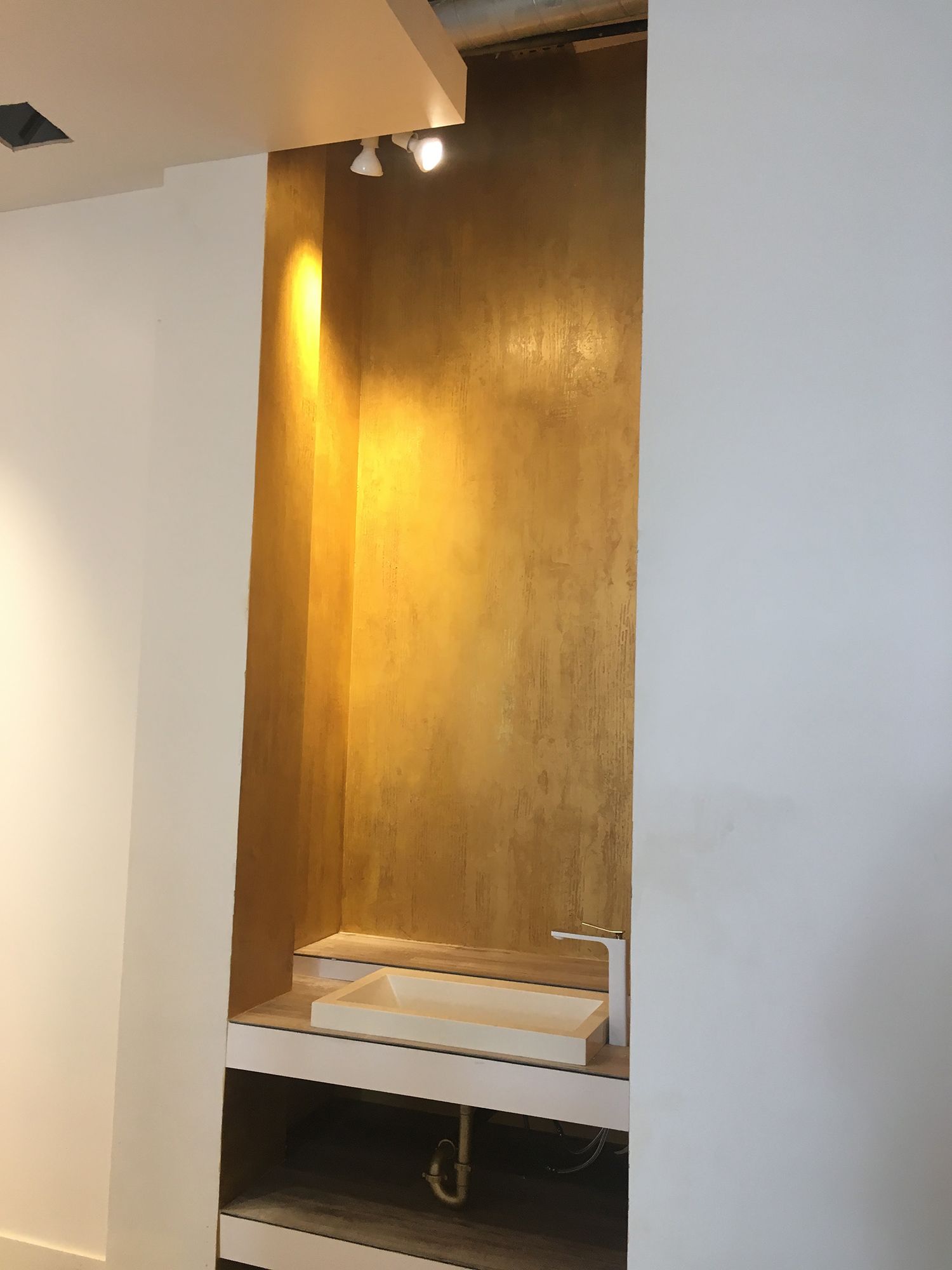 Recessed wall niche with gold-painted back, built-in sink, and spotlights. White walls.