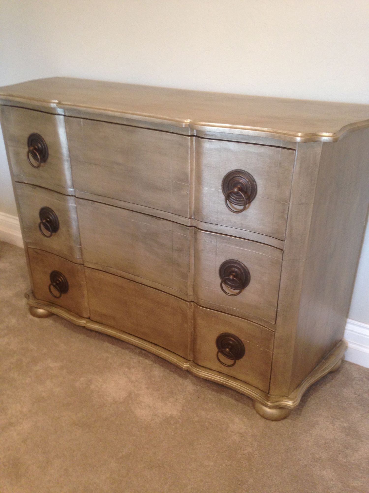 Silver chest of drawers with three drawers, round pulls, and a curved front, set on a beige carpet.