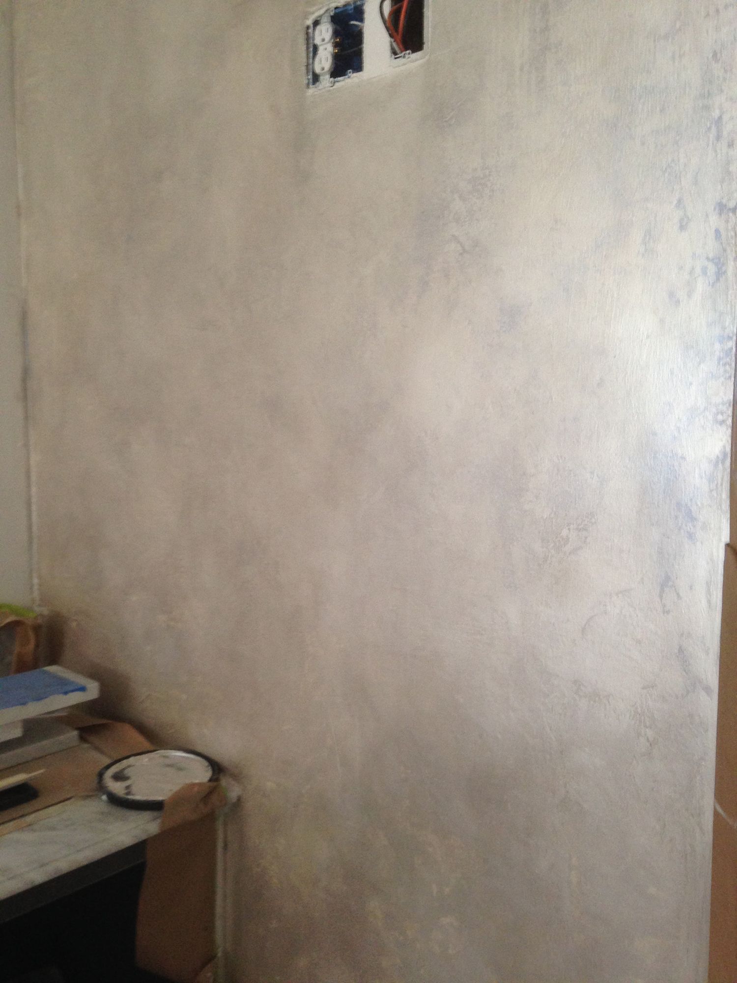 Textured, light-colored wall with an electrical outlet and a desk with items in the corner.