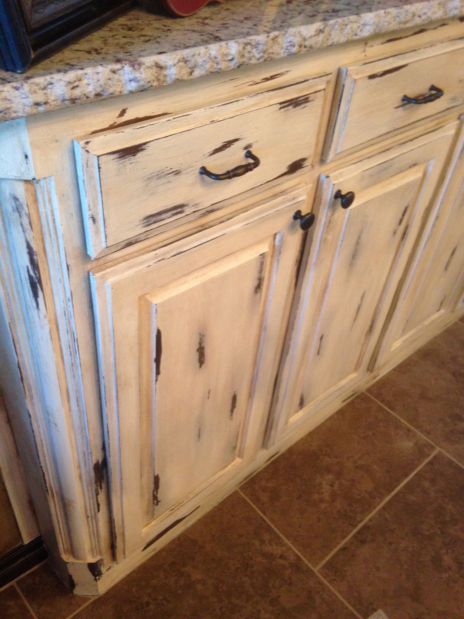 Cream-colored, distressed kitchen cabinets with dark brown accents and black hardware, set against a granite countertop.
