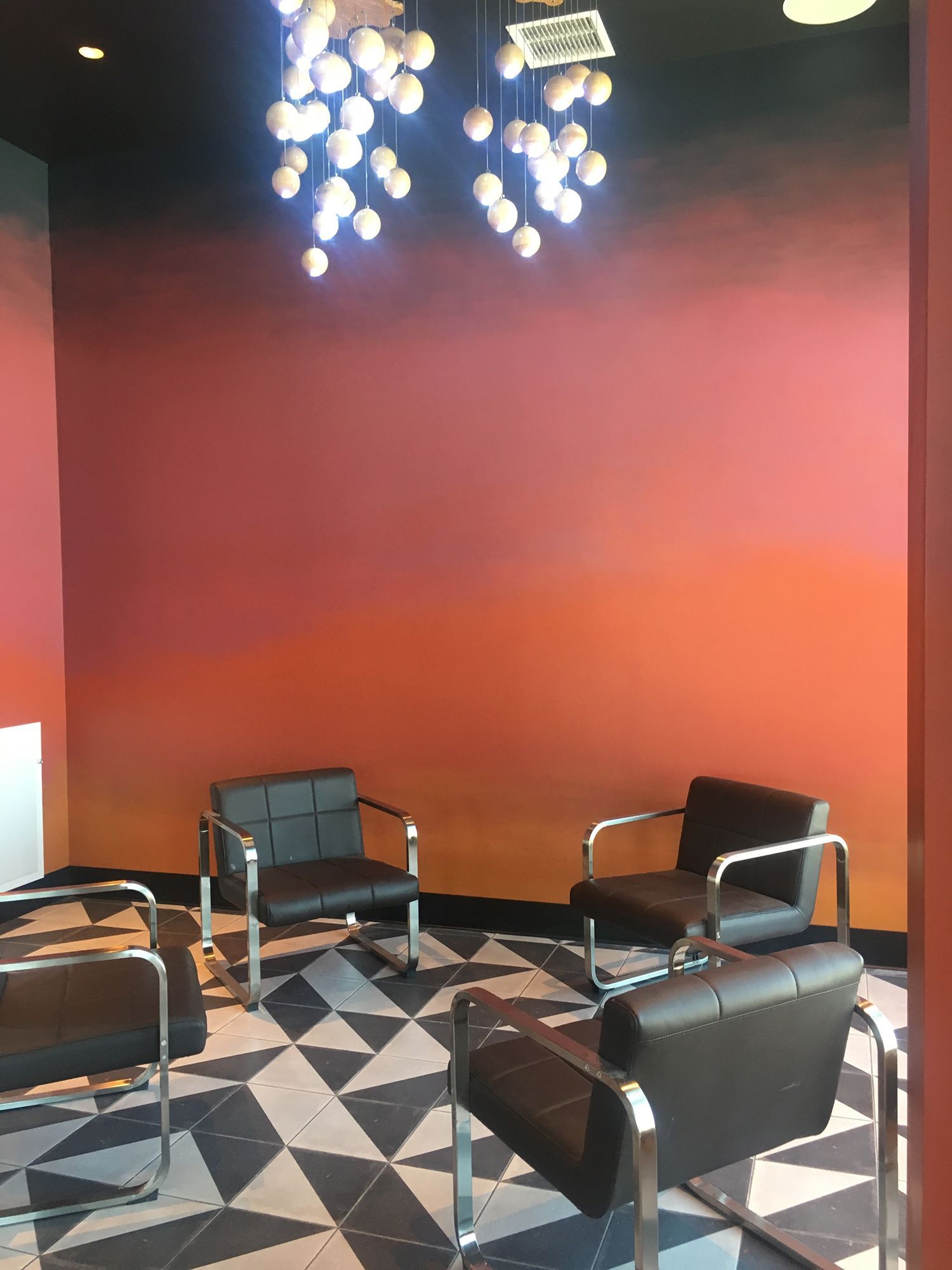 Four black chairs arranged in a circle on a patterned floor, with a gradient sunset-colored wall and hanging globe lights.