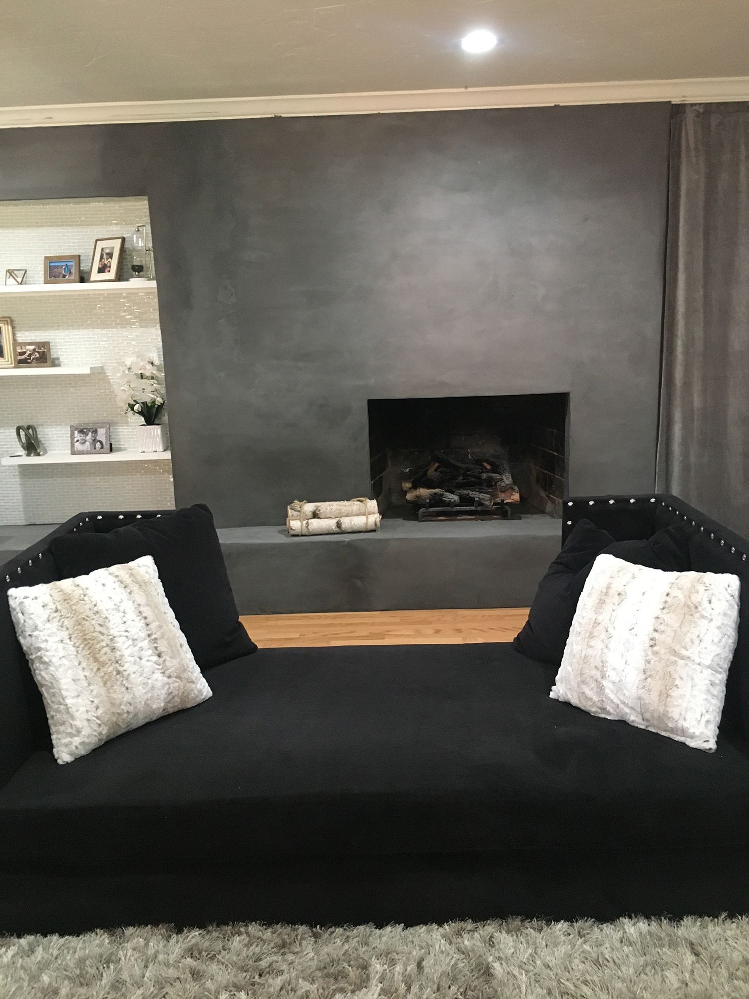 Black couch with white pillows in front of a gray fireplace and built-in white shelves.