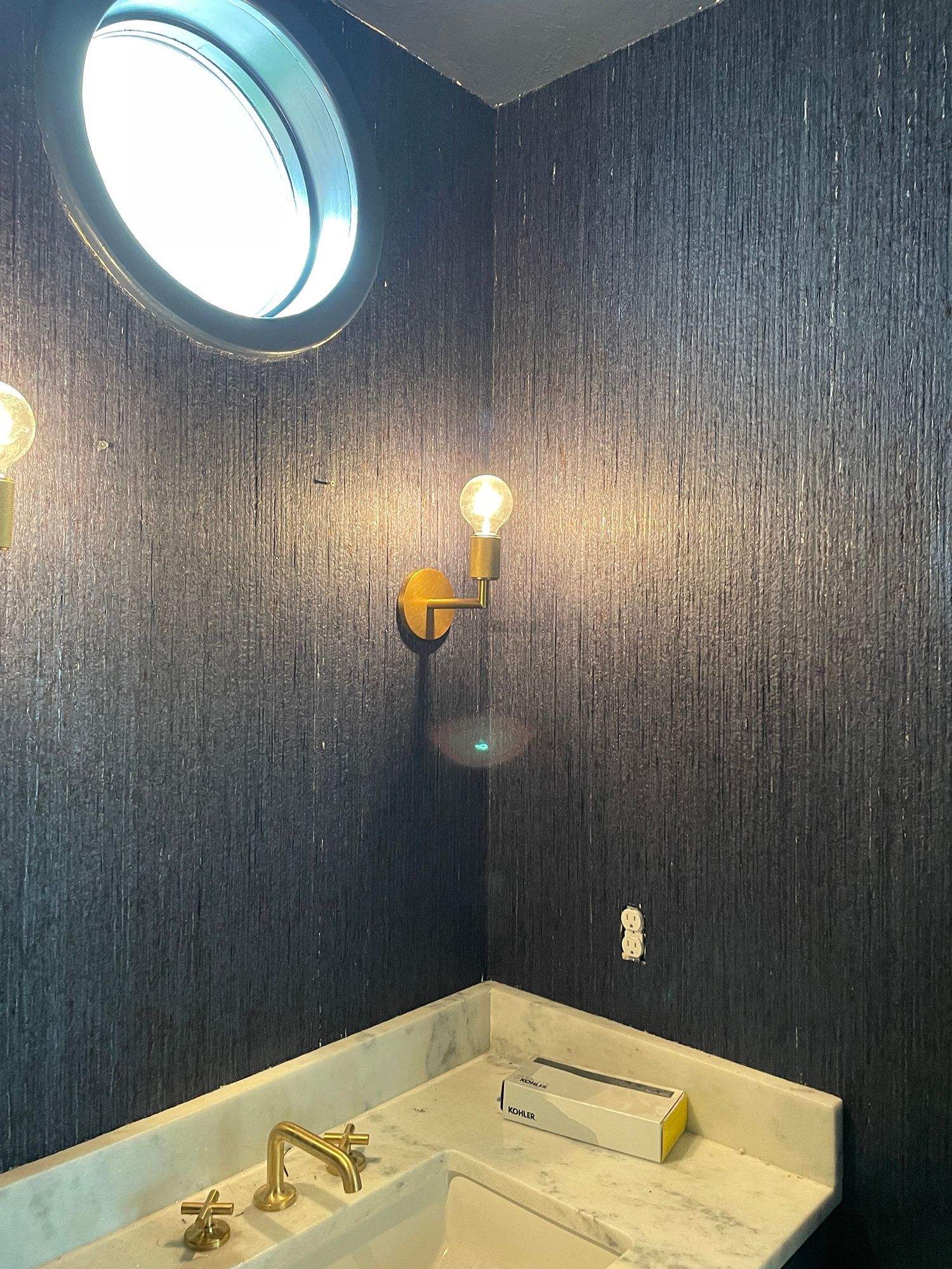 Small bathroom with dark, textured wallpaper, gold fixtures, and round window.