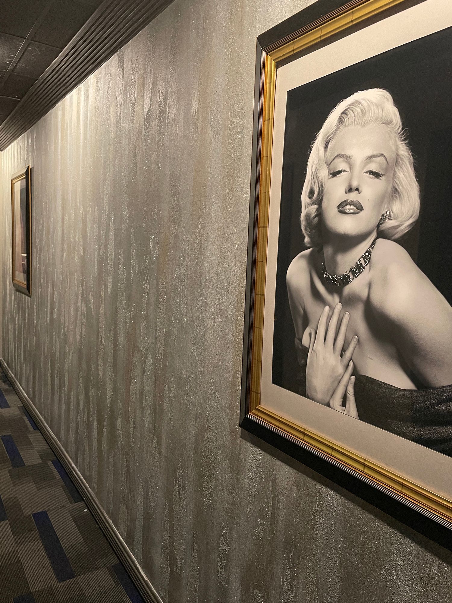 Hallway with Marilyn Monroe portrait in a gold frame. Walls are silver textured. Carpeted floor.