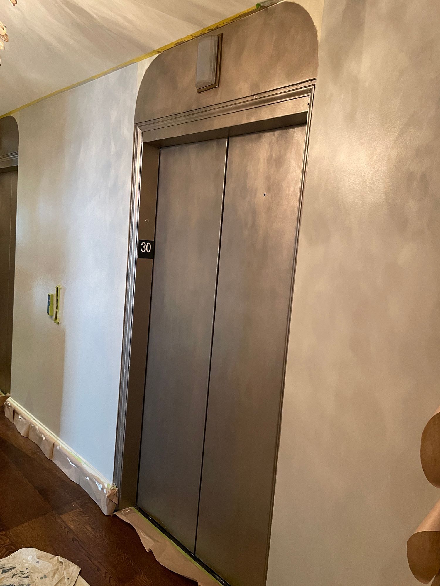 Elevator with metallic doors in a hallway with textured light gray walls and brown flooring.